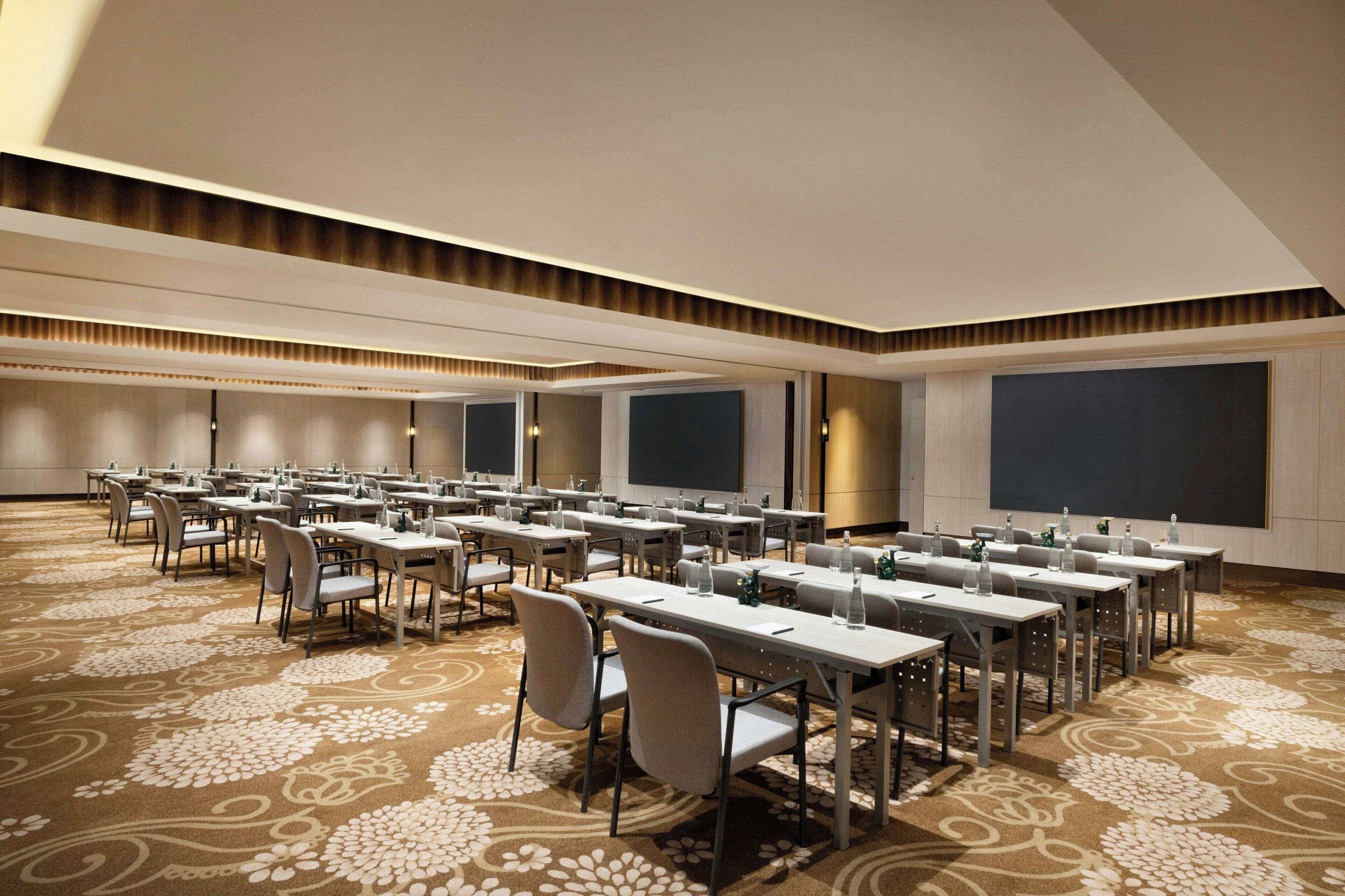 A conference room with rows of tables and chairs, large projection screens, and a patterned carpet.