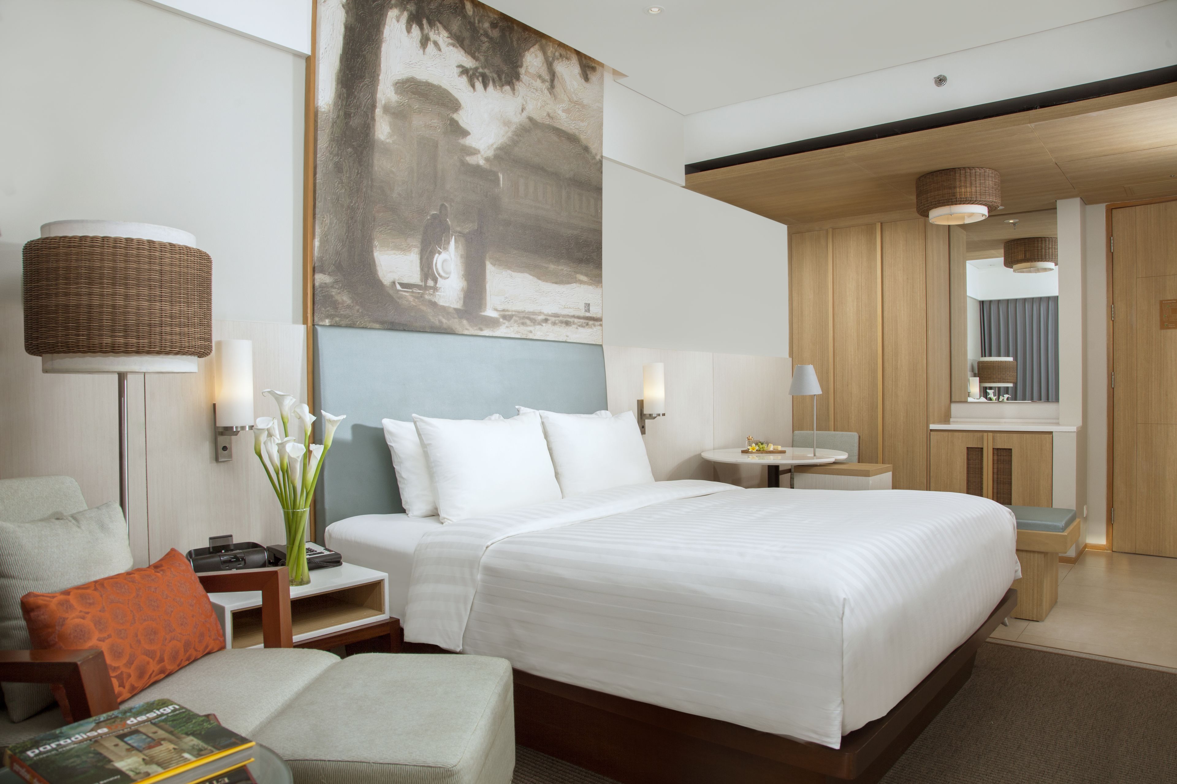 Una habitación de hotel moderna con una cama grande, una zona de estar con una silla y una mesita, un escritorio con lámpara, y un cuadro en la pared.