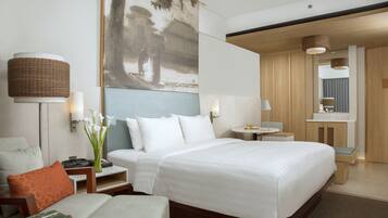 Una habitación de hotel moderna con una cama grande, una zona de estar con una silla y una mesita, un escritorio con lámpara, y un cuadro en la pared.
