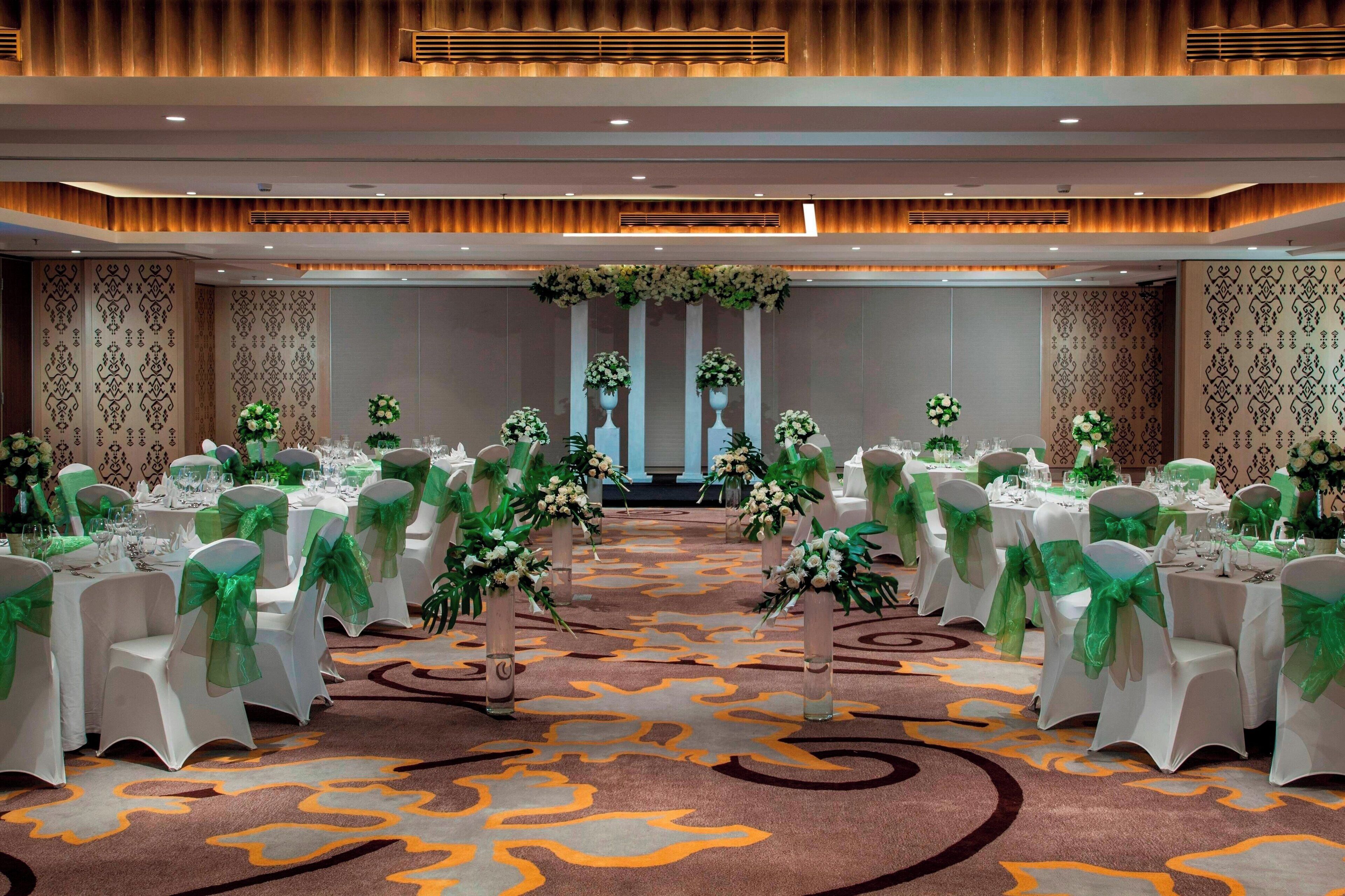 A banquet hall with rows of tables, each adorned with white tablecloths and green sashes, and decorated with floral centerpieces.
