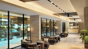 Un moderno vestíbulo de hotel con un mostrador de recepción alargado, zona de estar con sofás y sillones, ventanas amplias que ofrecen vistas a una piscina y un área exterior, y un ventilador de techo.