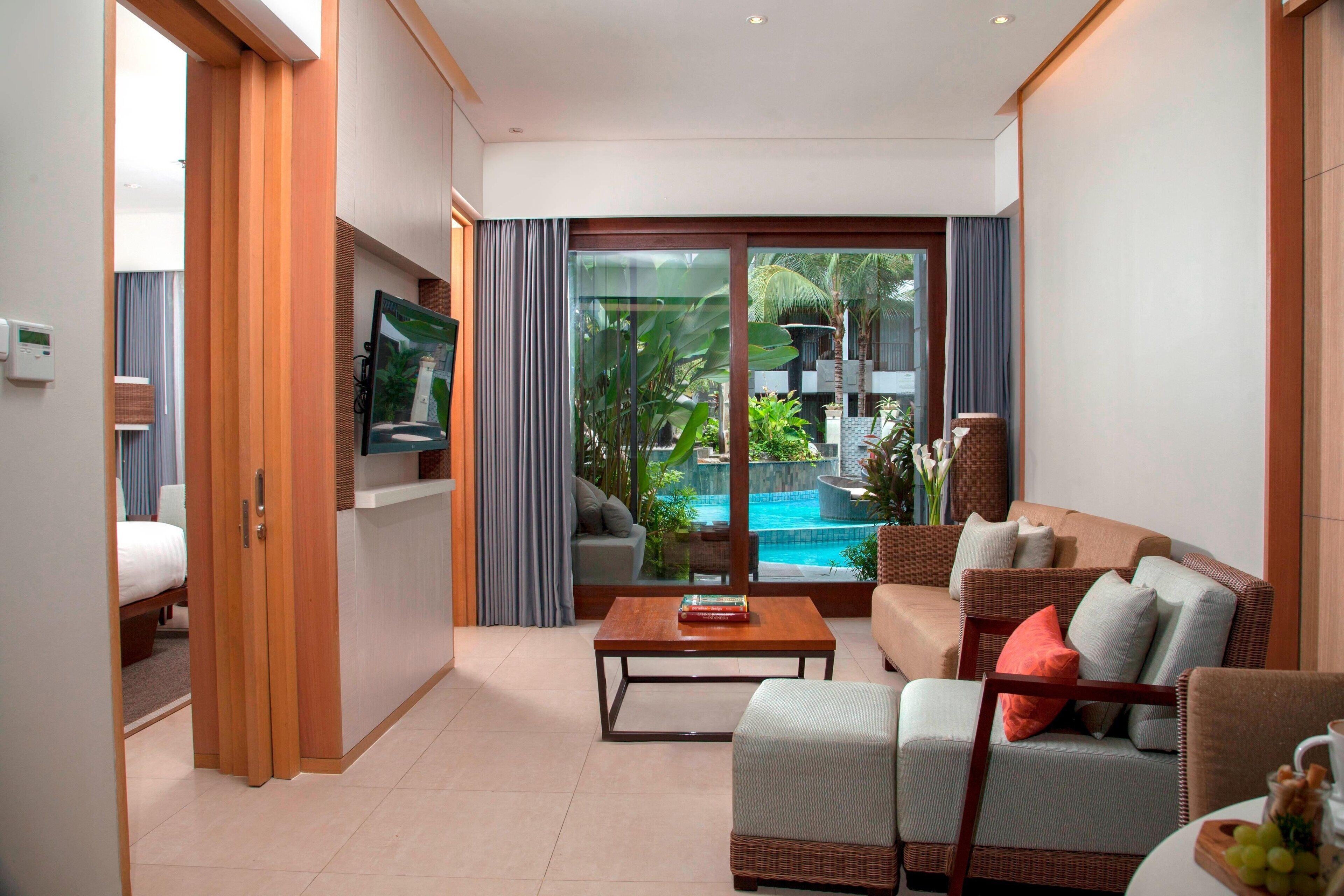 A modern living room with a sofa, a coffee table, and a TV. There is a sliding glass door leading to a pool area with tropical plants.