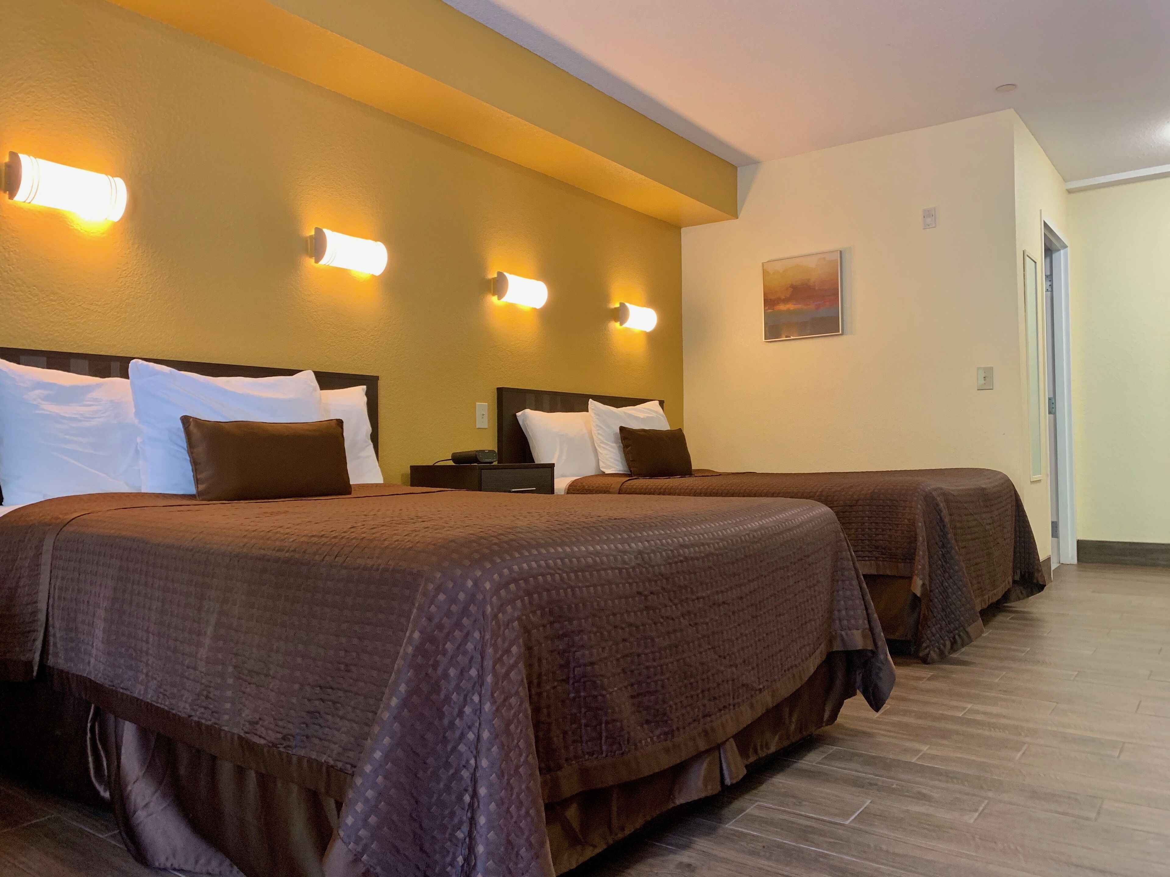 A hotel room with two beds, each with a brown bedspread and white pillows. There are wall-mounted lights on the left side of the room.