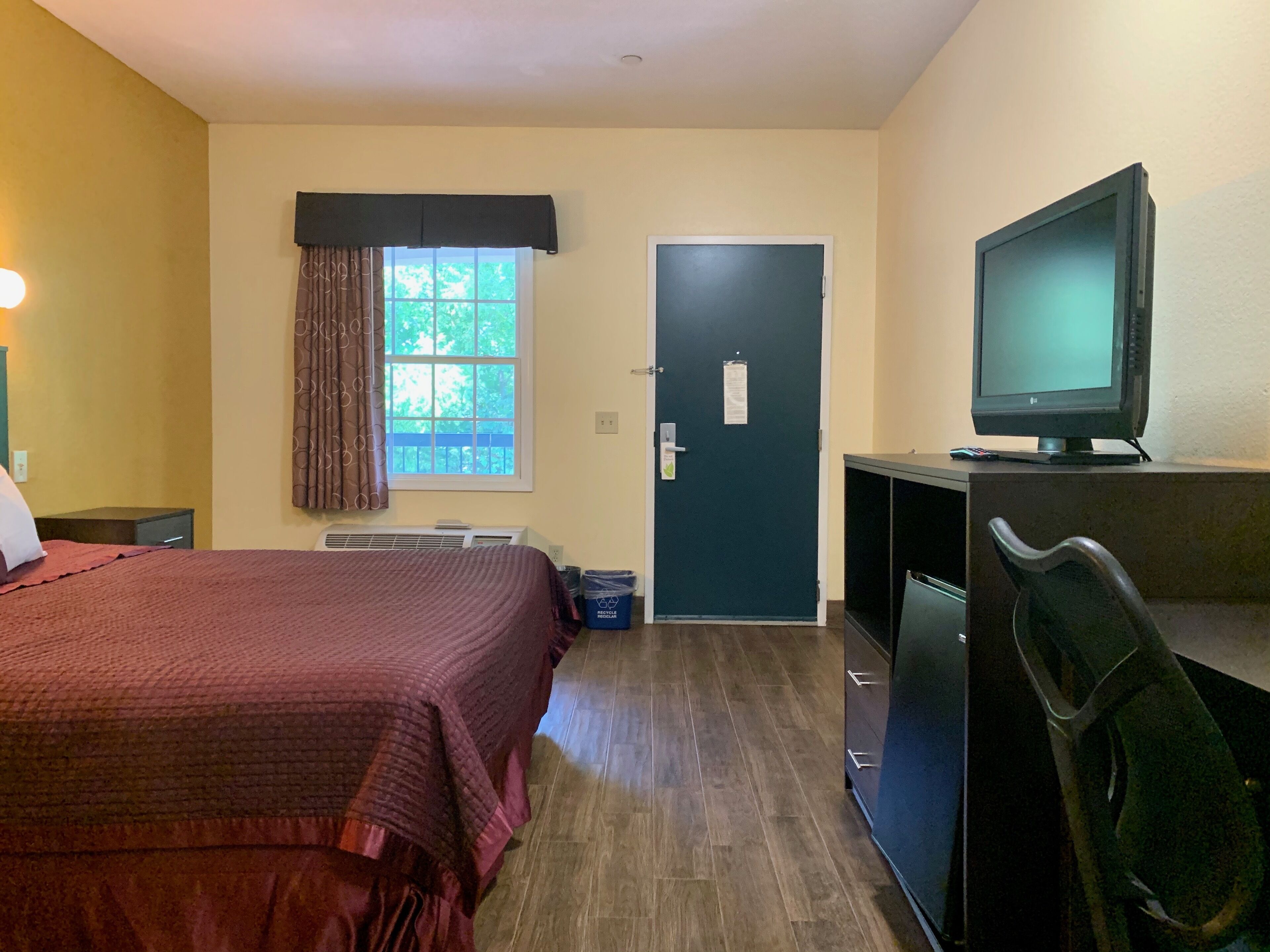 A hotel room with a bed, a desk with a computer, a chair, a window with curtains, and a door.