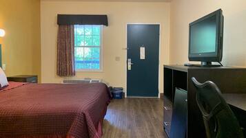 A hotel room with a bed, a desk with a computer, a chair, a window with curtains, and a door.