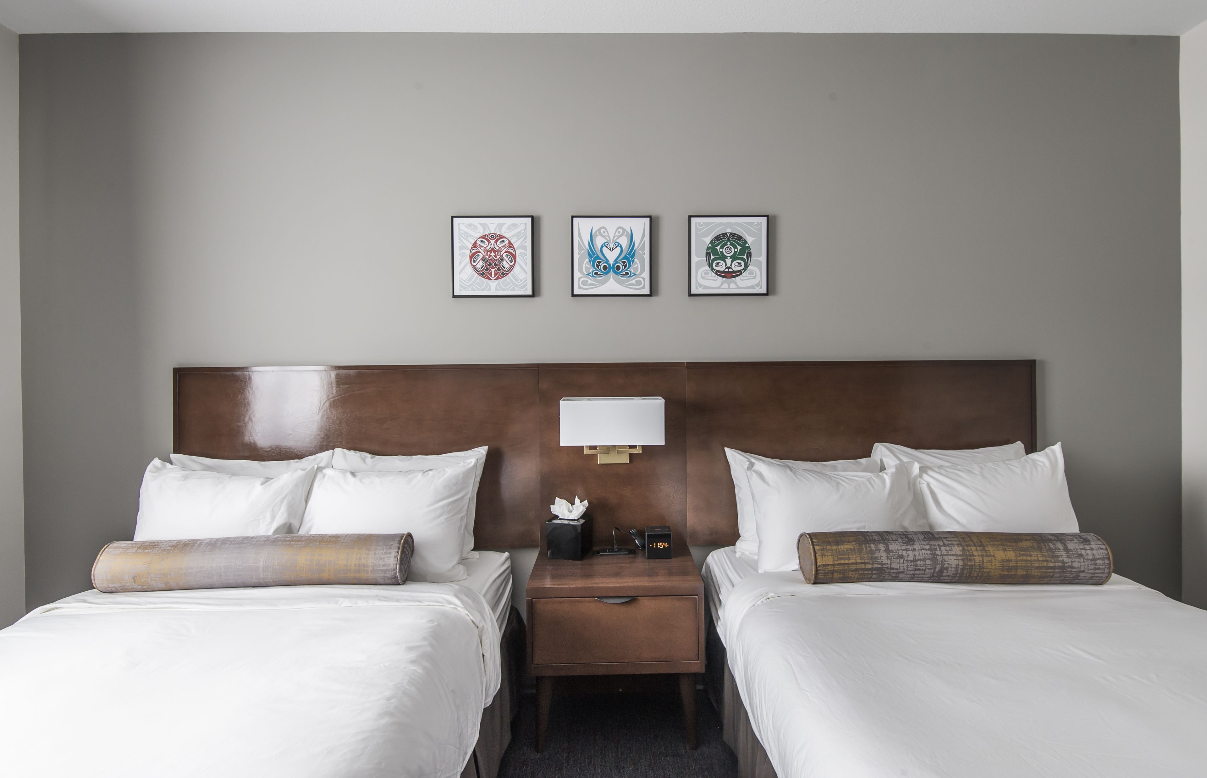 A hotel room with two beds, a nightstand with a lamp, and three framed artworks on the wall.