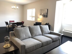 A modern living room with a grey sofa, a dining table with chairs, a chandelier, a lamp, and a painting on the wall. - Edgewater Hotel (Whitehorse)