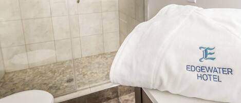Un bagno d'hotel con cabina doccia in vetro, lavandino bianco e un letto con cuscino che mostra il logo dell'hotel.