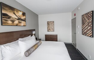Habitación de hotel con una cama grande, cabecero, mesita de noche, lámpara, mesita auxiliar de madera y dos obras de arte en las paredes.