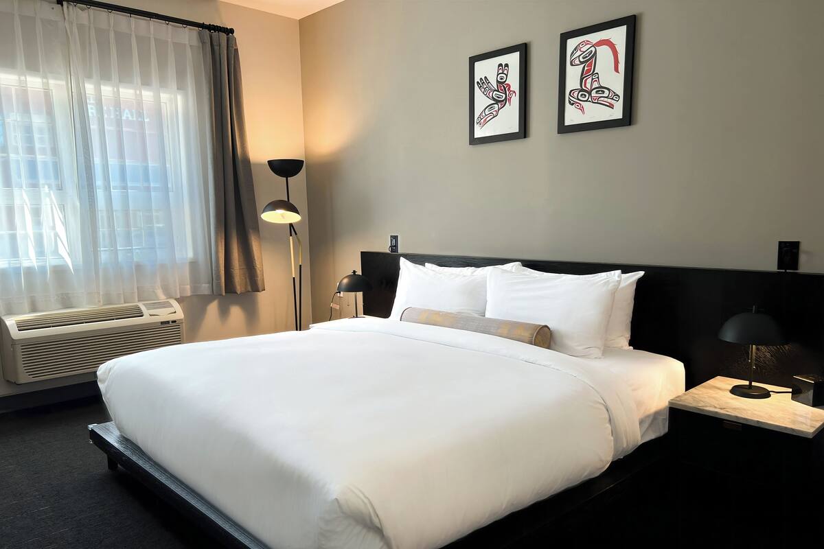 A hotel room with a large bed, a bedside table with a lamp, a wall-mounted air conditioner, and two framed artworks above the bed.