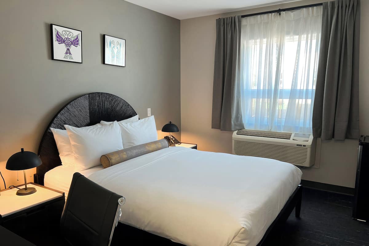 A hotel room with a large bed, a desk, two framed artworks, a window with curtains, and an air conditioning unit.