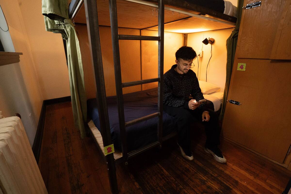 Une chambre avec un lit superposé, une personne assise sur le lit du bas, un plancher en bois et une porte avec une poignée.