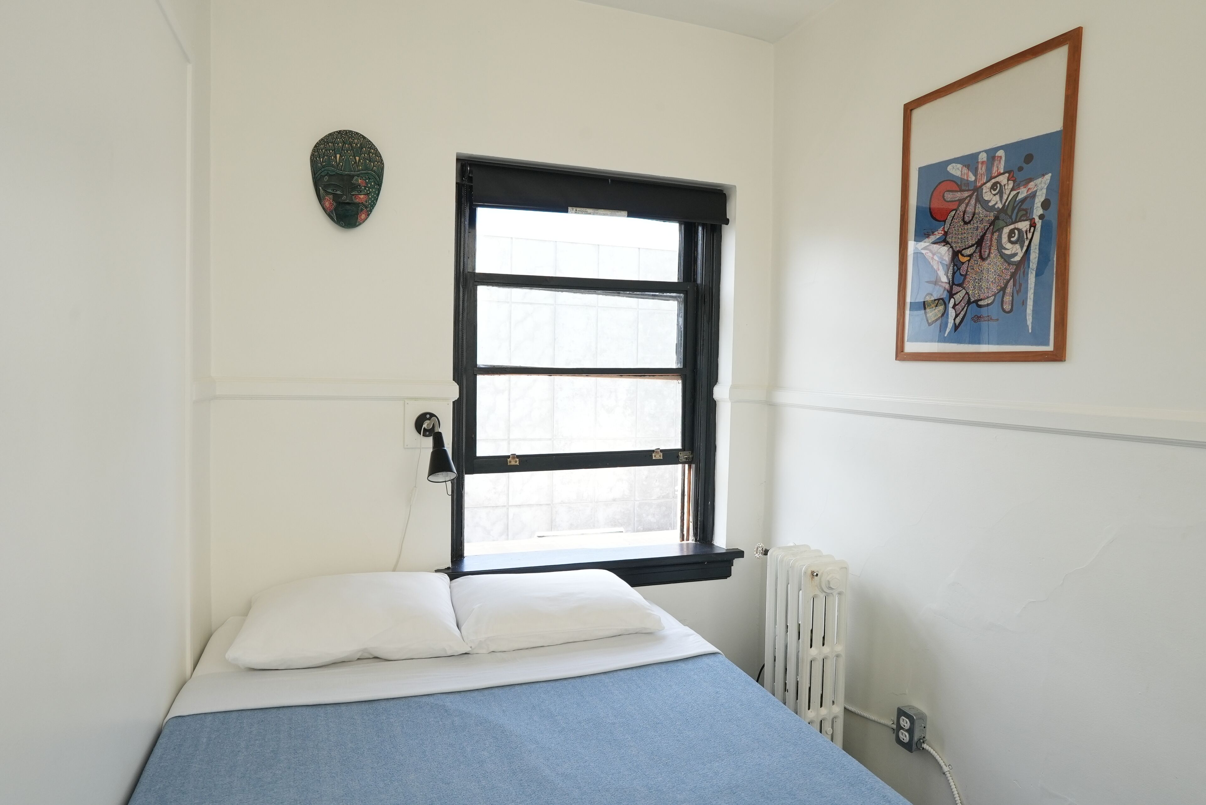 A bedroom with a single bed, a window with black frames, a radiator, and a wall-mounted artwork.