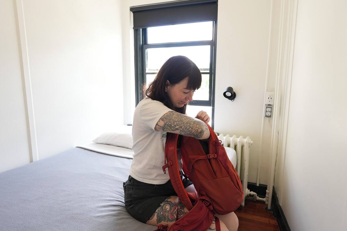 Une personne est assise sur un lit dans une pièce avec une fenêtre, et elle tient un sac à dos rouge.