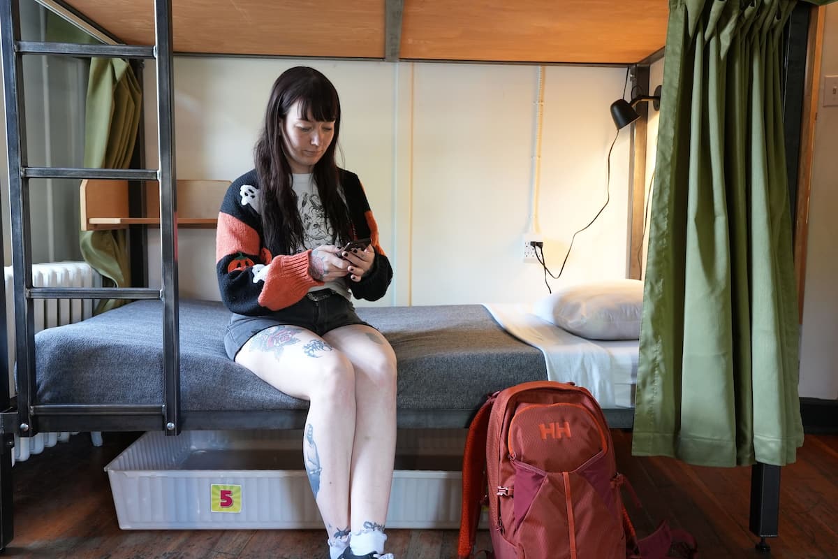 Un lit superposé avec un matelas, une échelle, un sac à dos rouge et une lampe fixée au mur.