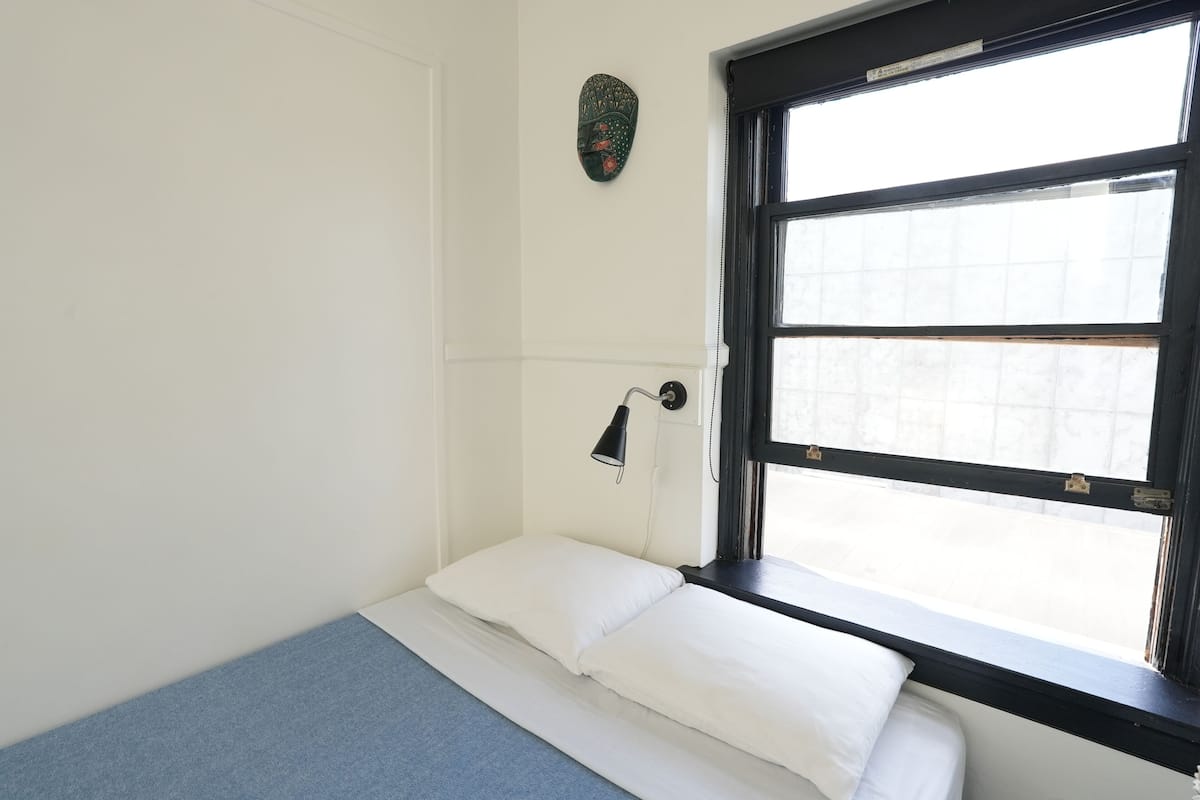 Une petite chambre avec un lit simple, une fenêtre, une lampe fixée au mur et un objet décoratif au-dessus du lit.