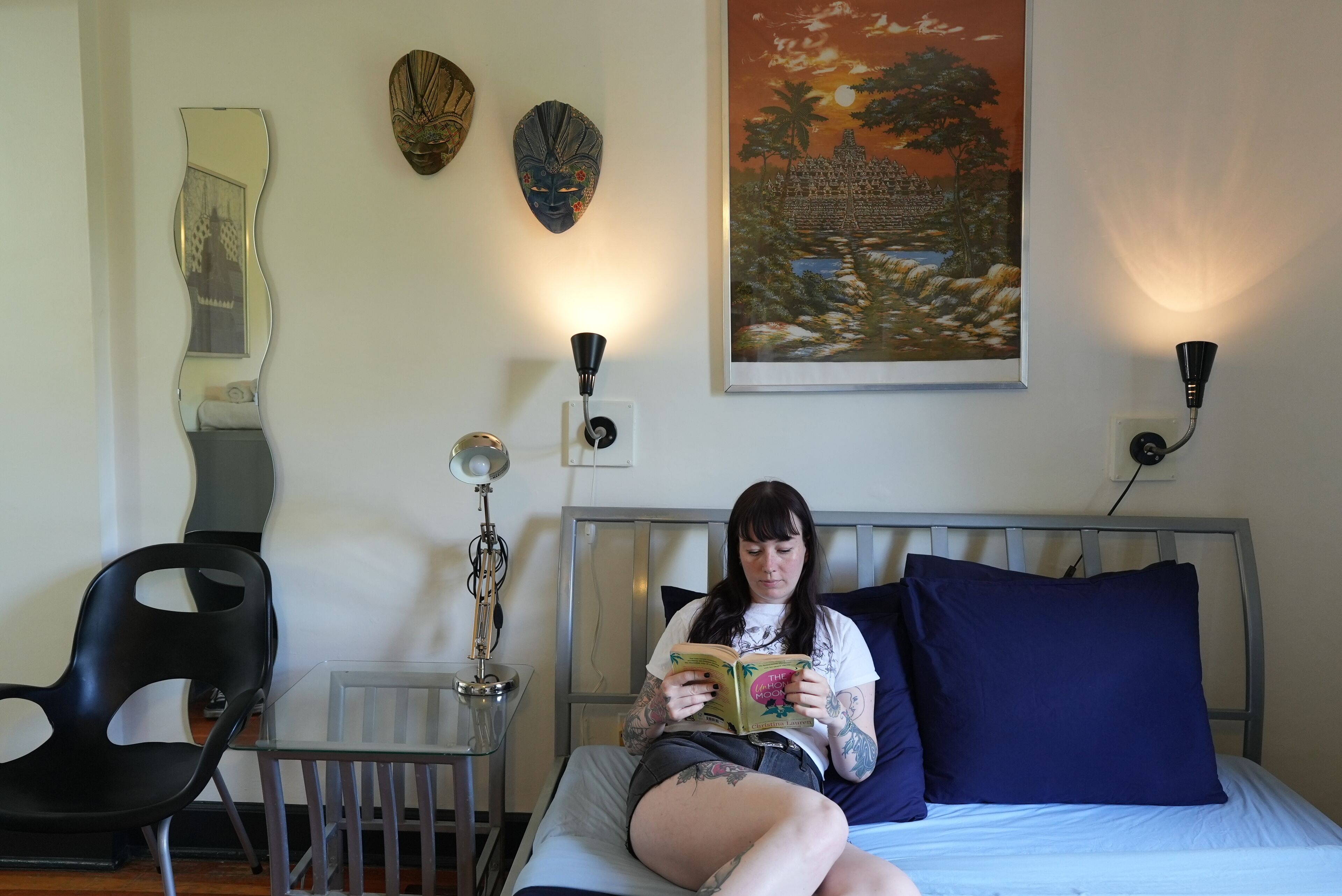 Une personne est assise sur un lit, en train de lire un livre dans une pièce où se trouvent un miroir, deux masques accrochés au mur et un tableau représentant un temple.