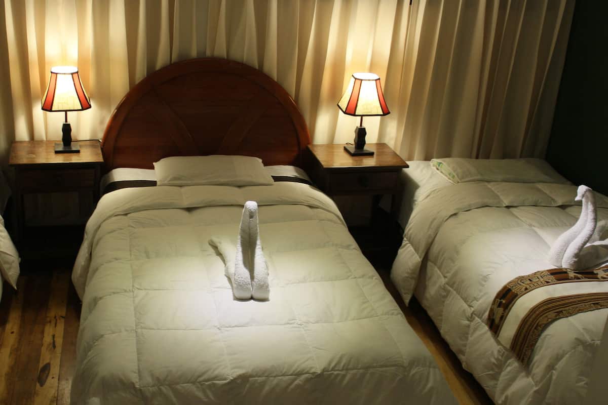 Une chambre d’hôtel avec trois lits, chacun agrémenté d’une serviette en forme de cygne. Il y a deux tables de chevet avec des lampes, et la chambre est dotée d’une tête de lit en bois.
