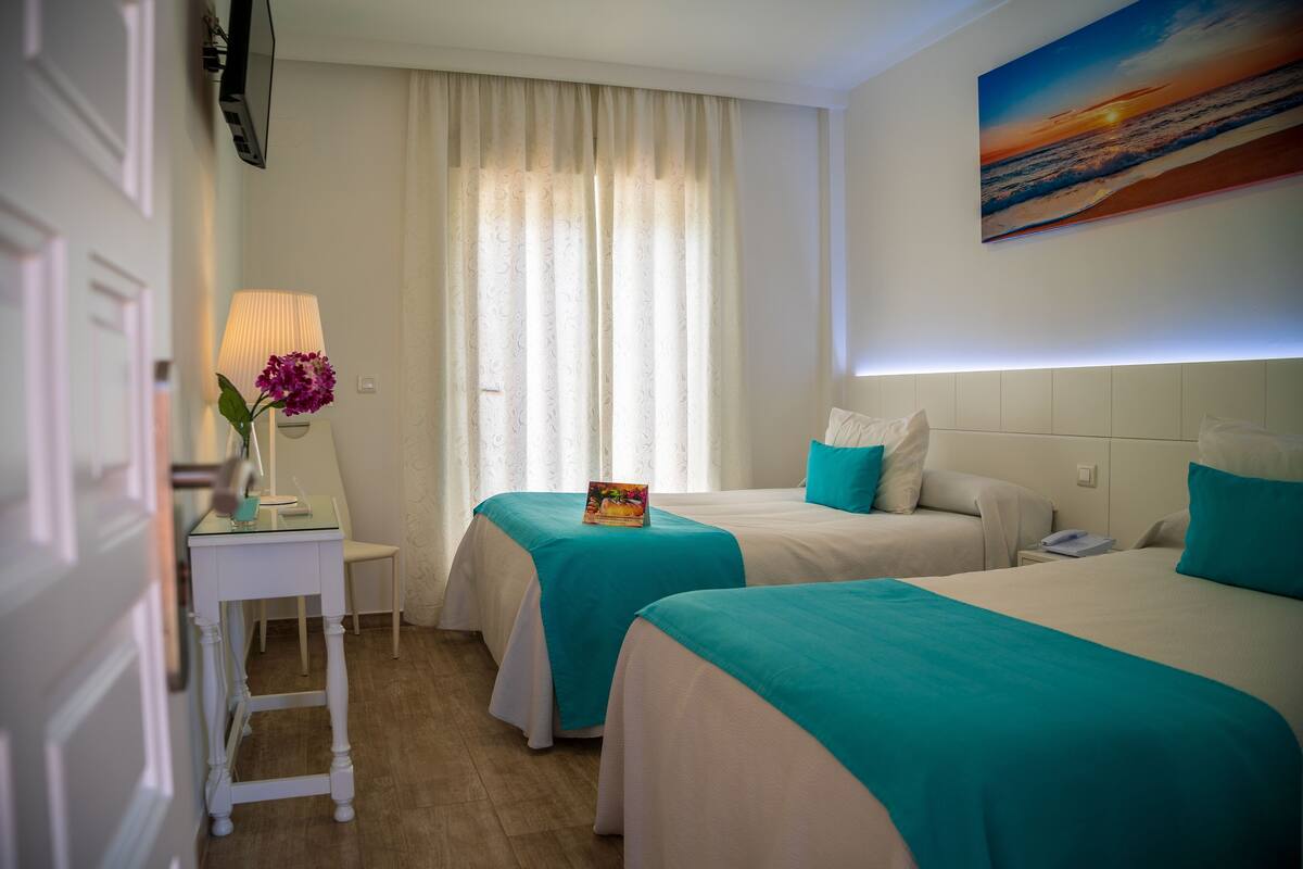 Habitación de hotel con dos camas, un escritorio con lámpara, una silla y un cuadro en la pared.