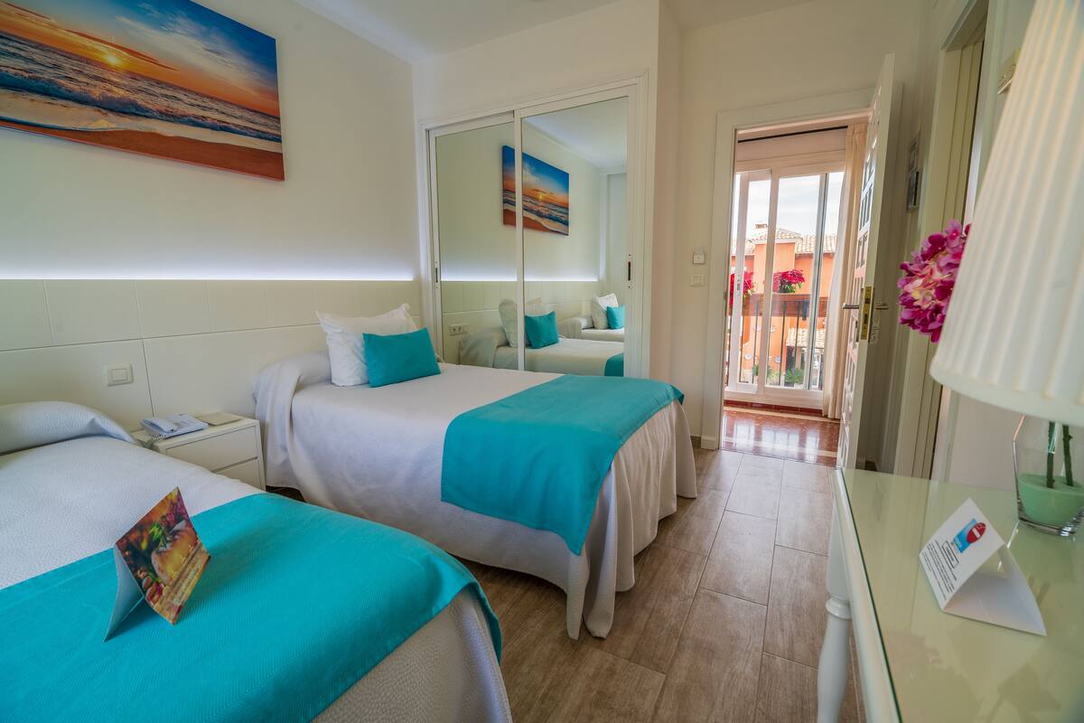 Habitación de hotel con dos camas, una mesita de noche, un escritorio con lámpara y vistas al balcón exterior.