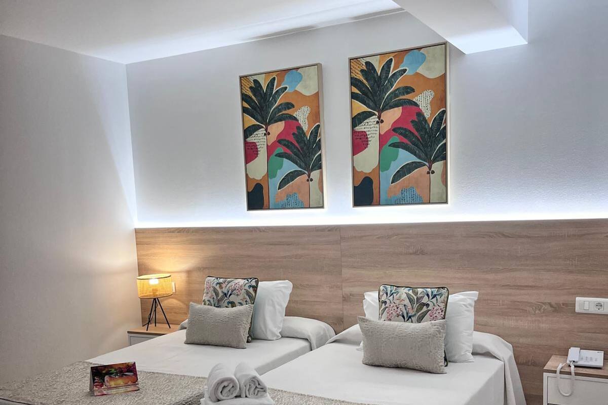 Habitación de hotel con dos camas, una mesita de noche, una lámpara y dos cuadros en la pared.