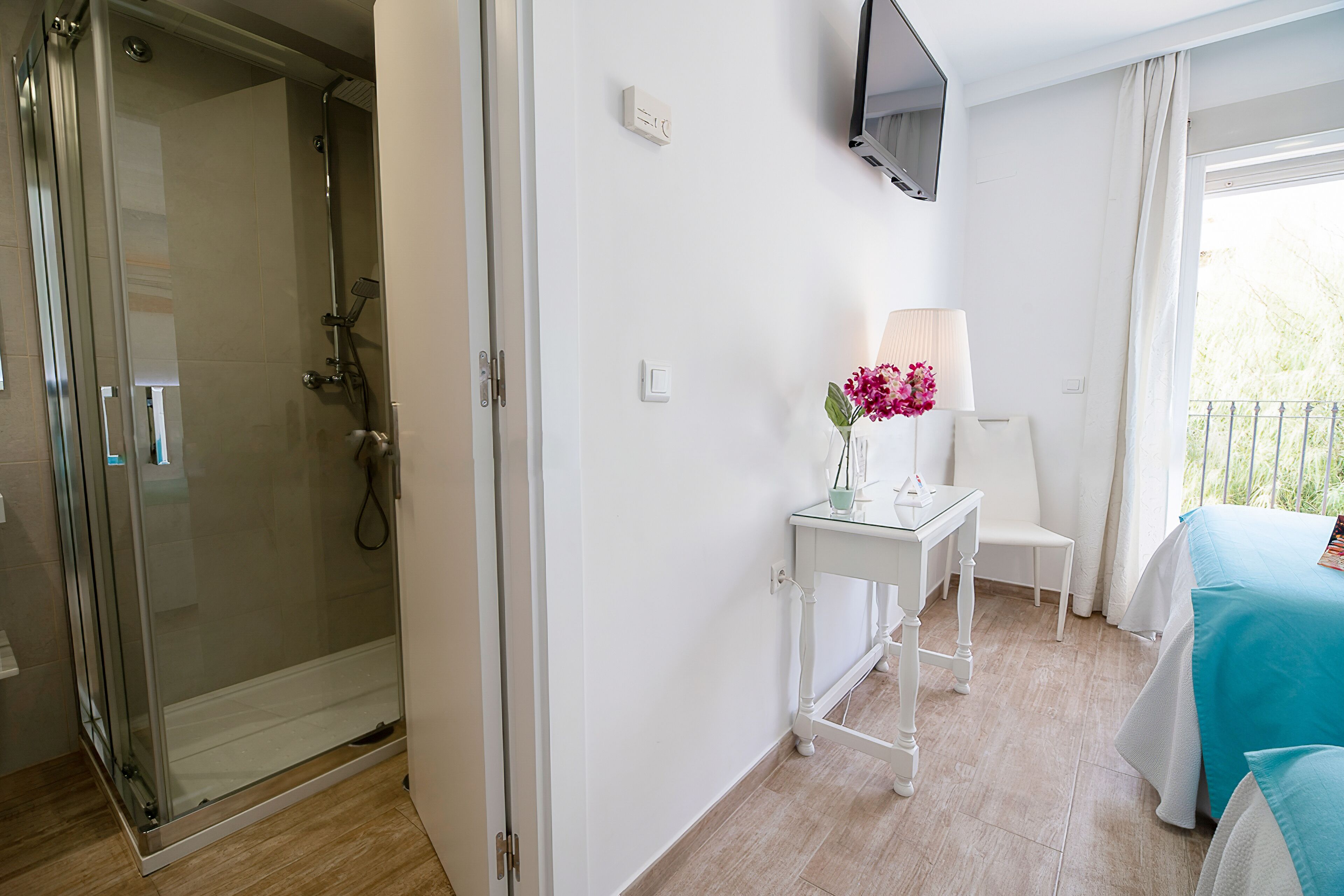 Une chambre d’hôtel avec une douche en verre, une petite table avec une lampe et des fleurs, un lit recouvert d’une couverture bleue et un téléviseur fixé au mur.
