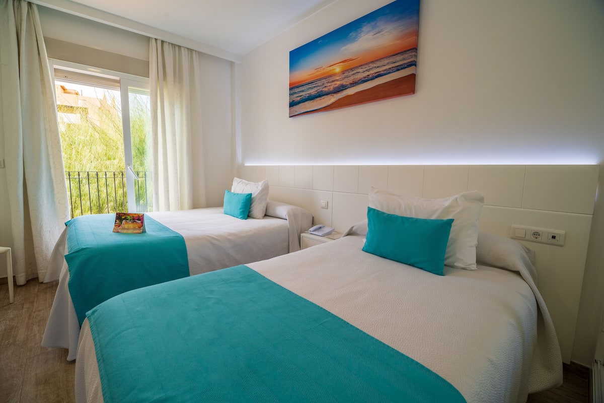 Habitación de hotel con dos camas, cada una con cubrecama color turquesa y almohadas blancas, un cuadro de un atardecer en la pared y una ventana con cortinas.