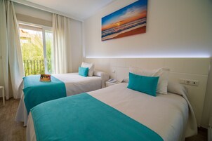 Habitación de hotel con dos camas, cada una con cubrecama color turquesa y almohadas blancas, un cuadro de un atardecer en la pared y una ventana con cortinas.