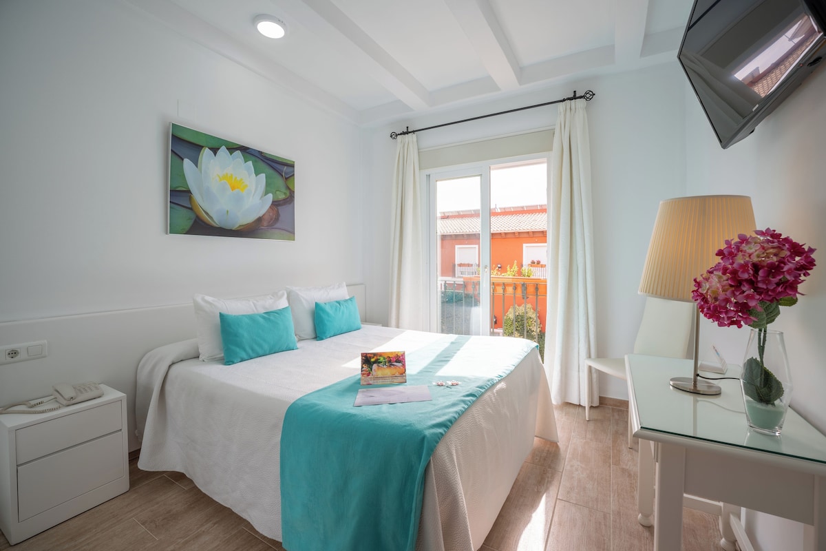 A hotel room with a bed, a bedside table with a lamp, a TV mounted on the wall, and a window with curtains.