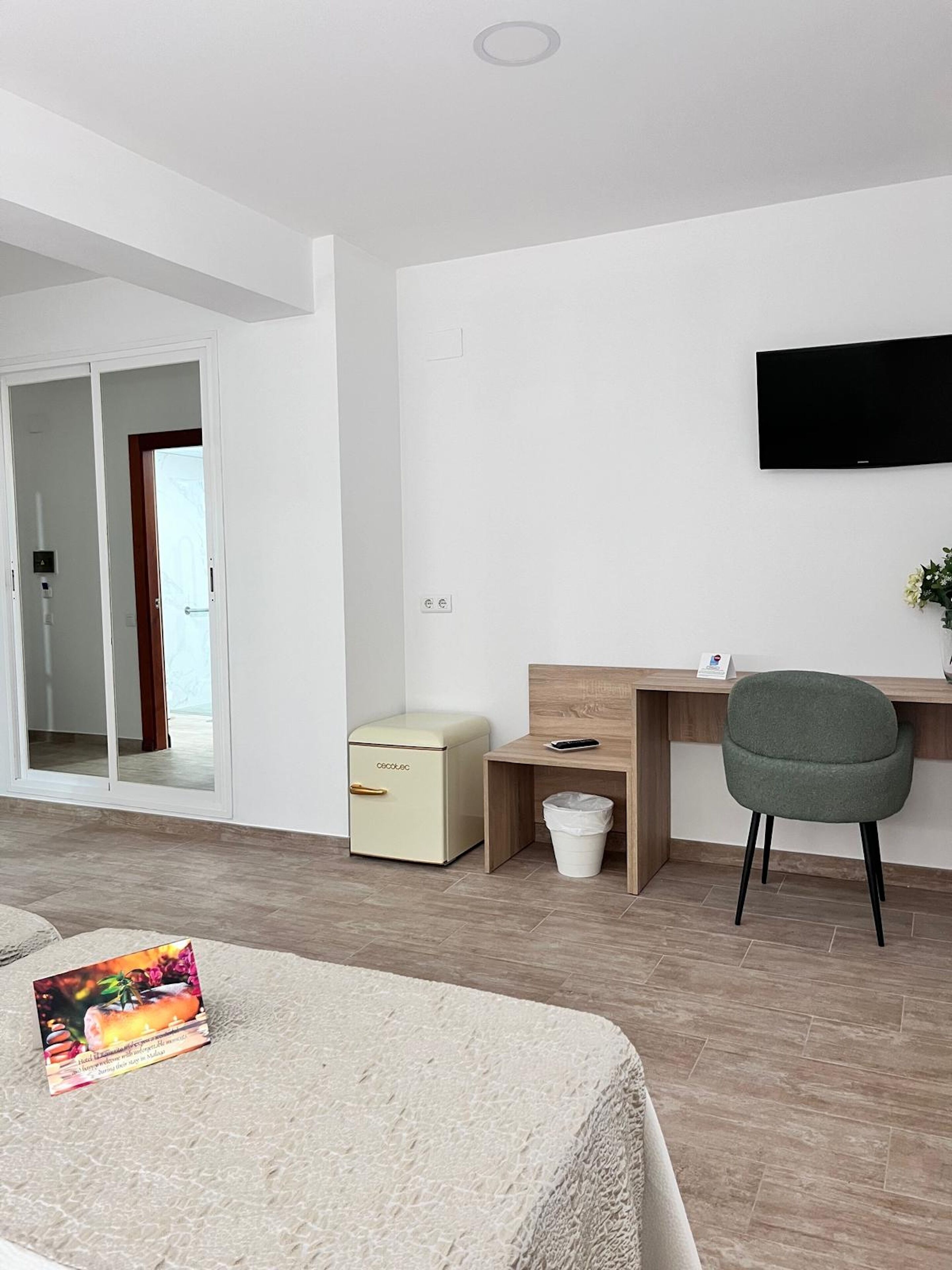 Une chambre d’hôtel moderne comprenant un lit, un bureau avec une chaise, un téléviseur fixé au mur, un petit meuble et une poubelle.