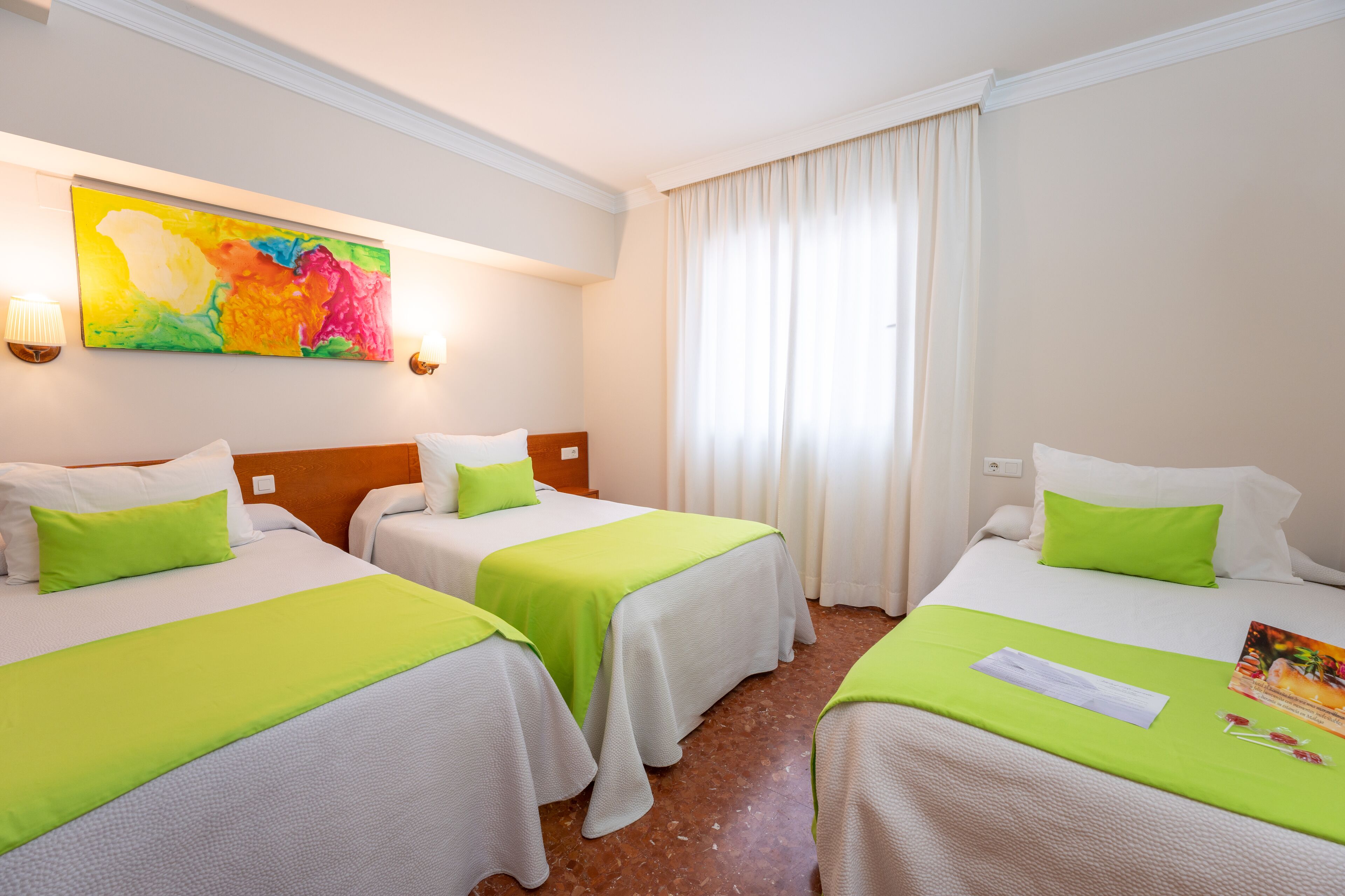 Habitación de hotel con dos camas, cada una con cubrecama verde y almohadas blancas. En la pared, un cuadro abstracto de colores; una ventana con cortinas blancas y una lámpara en la mesita de noche.