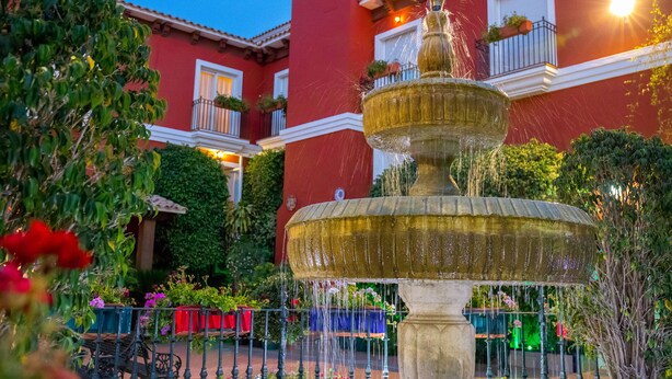 A multi-tiered fountain in a courtyard with a red building in the background. - Hotel Romerito (Málaga)