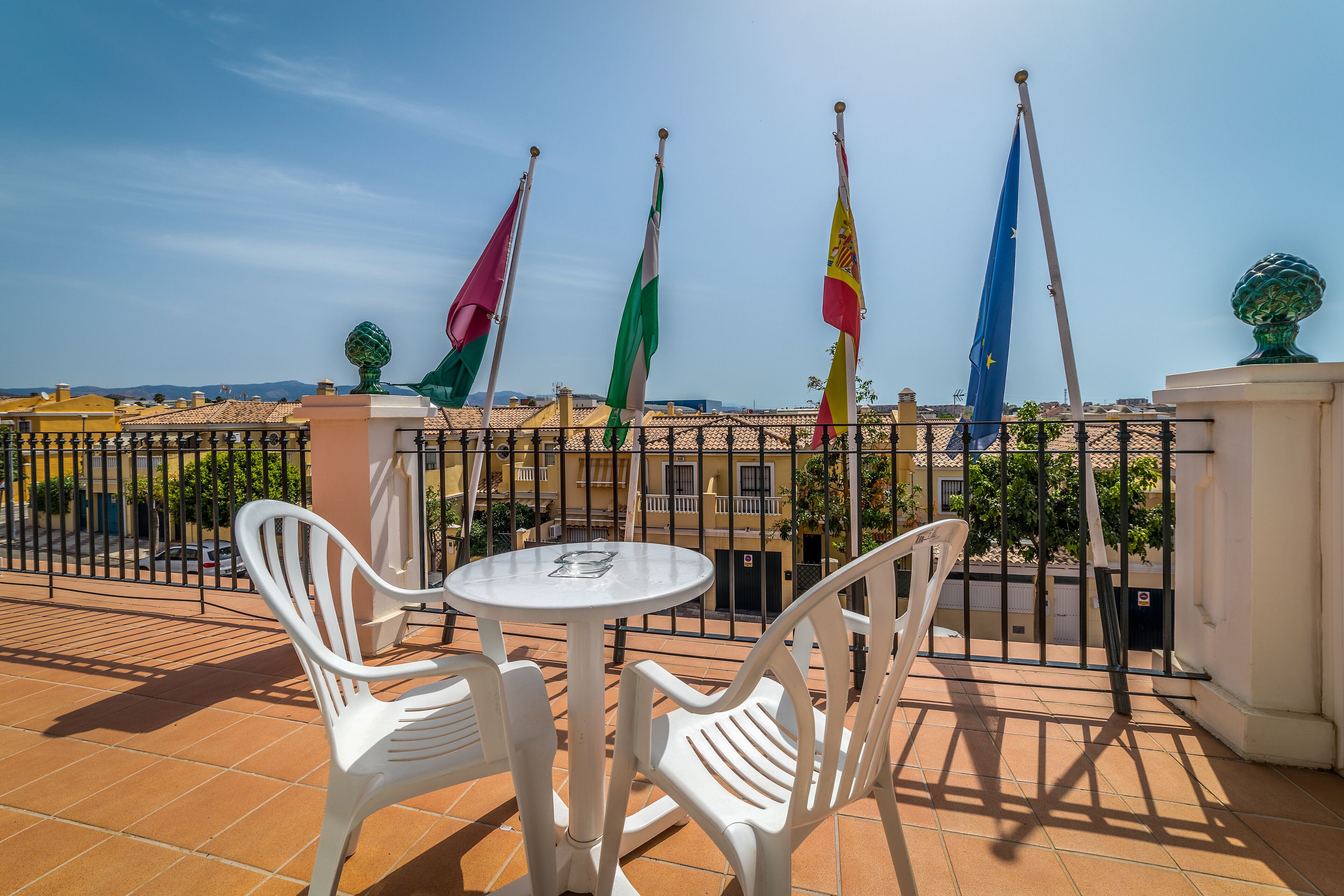 Une terrasse avec des chaises et une table en plastique blanc, entourée d’une clôture en métal ornée de drapeaux.
