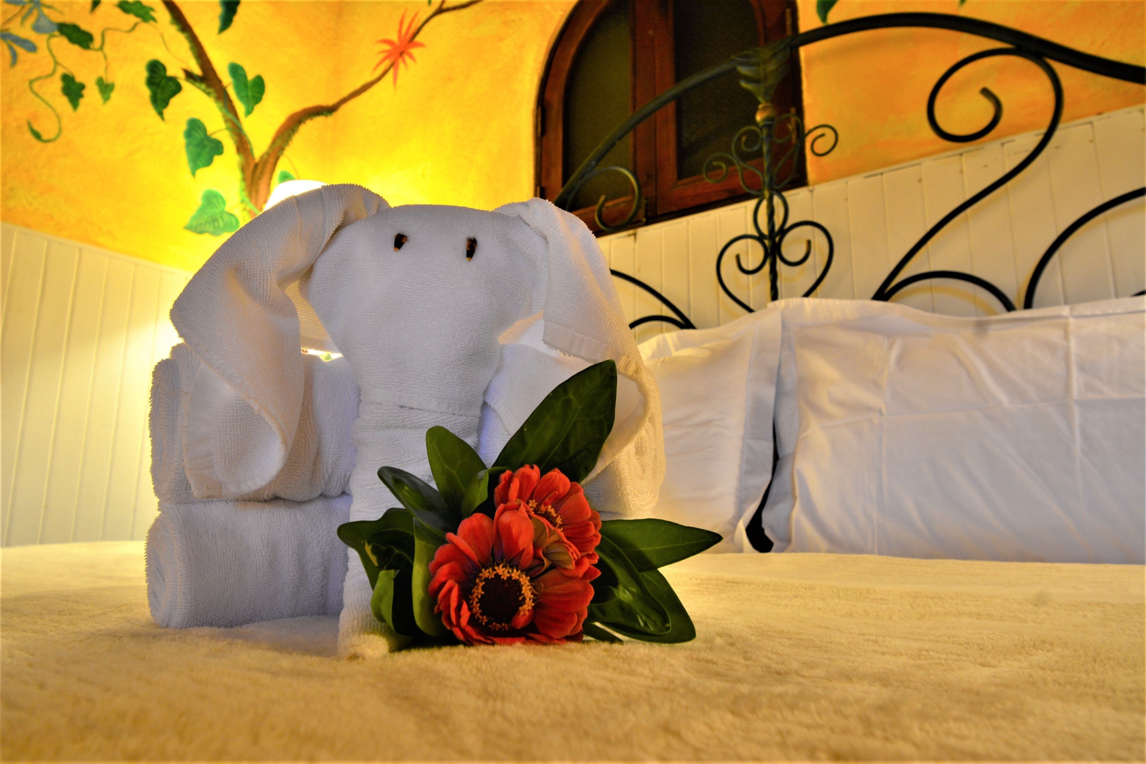 A bed with a white towel folded into an elephant shape, a red flower, and a decorative wall with a tree and leaves.