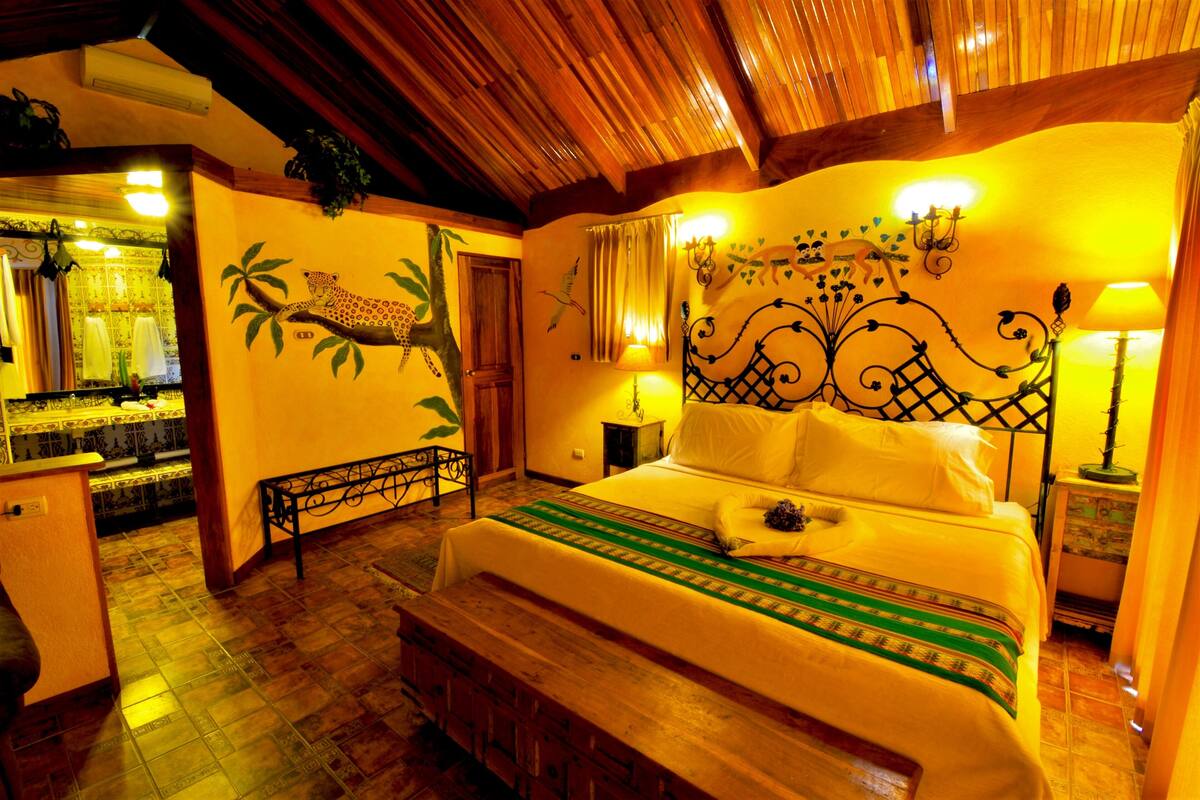 Un dormitorio con cabecera de madera, cama con manta verde y blanca a rayas, y mesita de noche con lámpara.