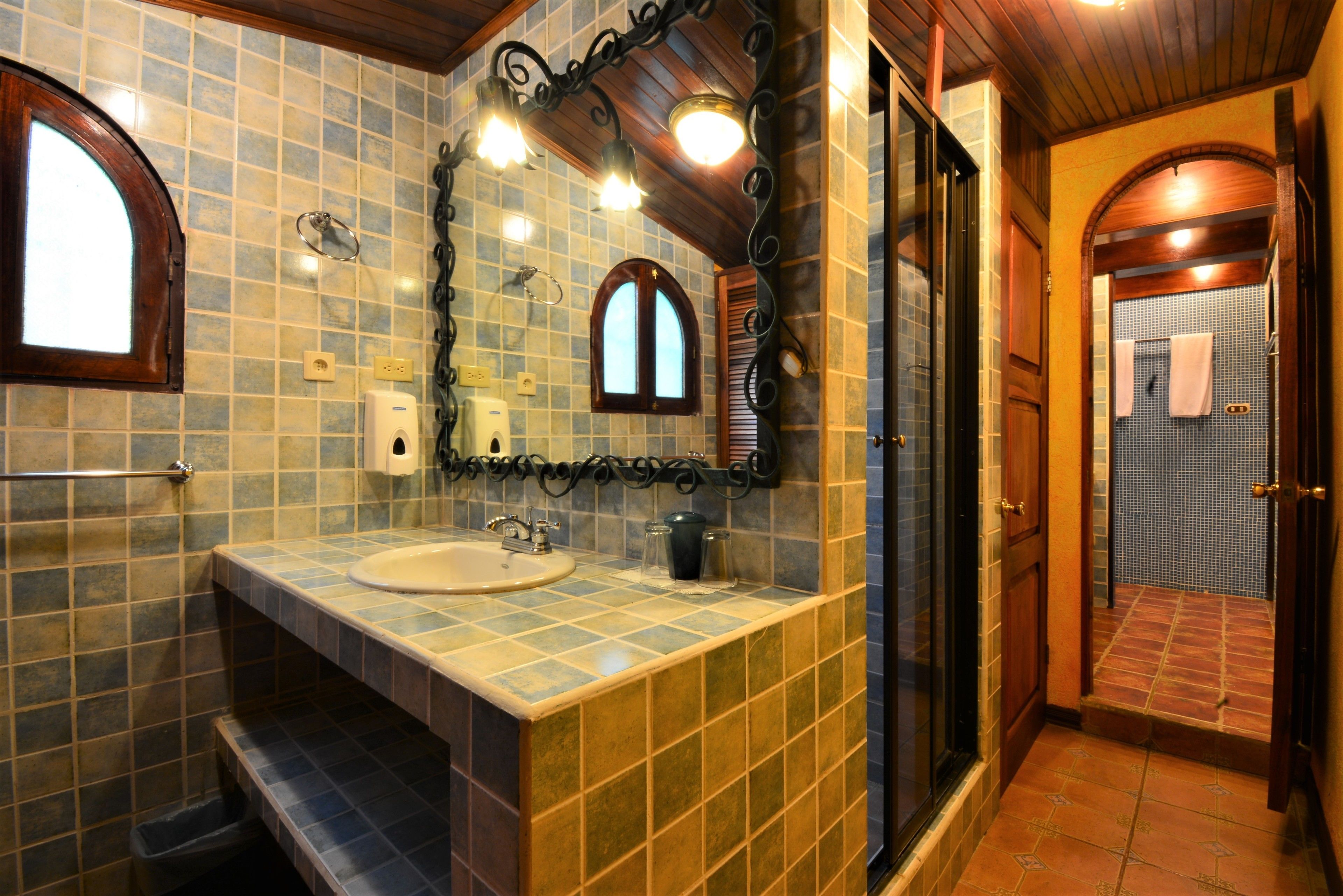 A bathroom with a tiled wall, a sink, a mirror, and a shower area.