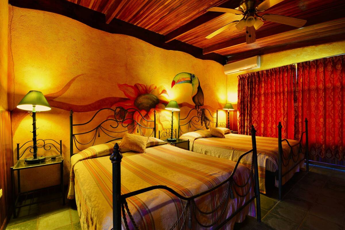 Habitación con dos camas, ventilador de techo y una ventana grande con cortinas rojas.