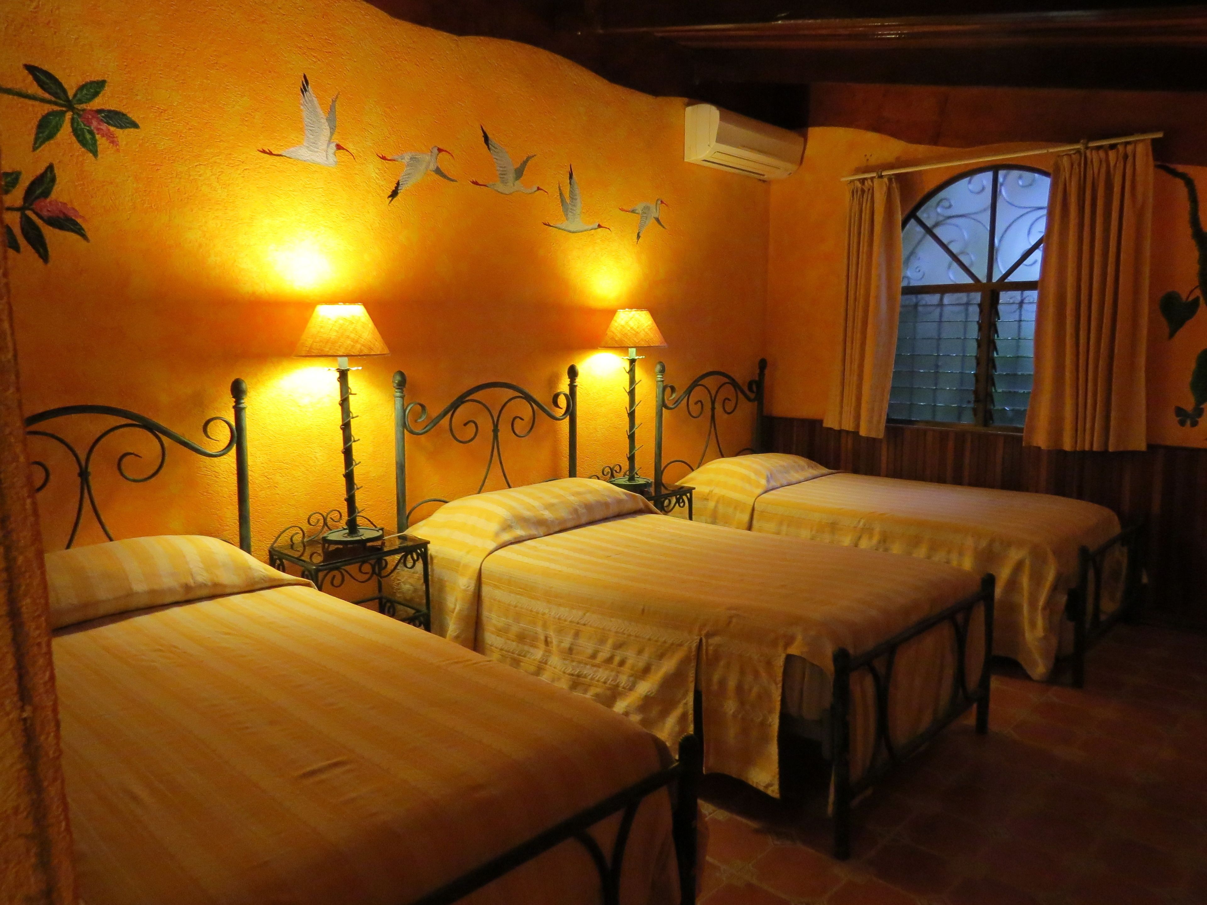 A room with four beds, each with a lamp and a small table beside it. There is a window with curtains and a decorative wall with birds and leaves.