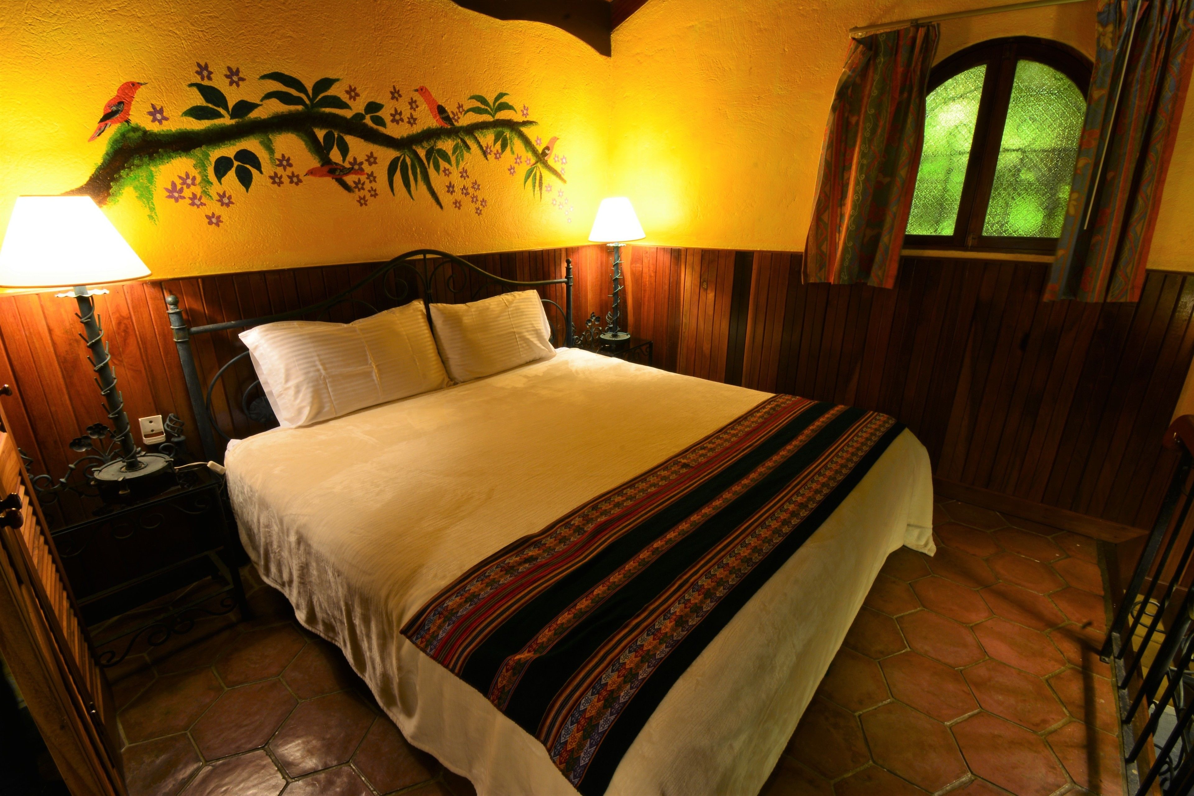 Uma cama com colcha branca e manta colorida, duas luminárias de cabeceira, um mural na parede com uma árvore e pássaros, e uma janela com cortinas.