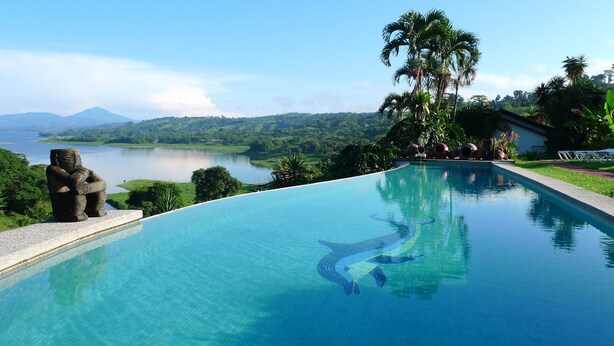 A pool with clear blue water, a stone statue, and a view of a lake and mountains. - La Mansion Inn Arenal (Arenal)