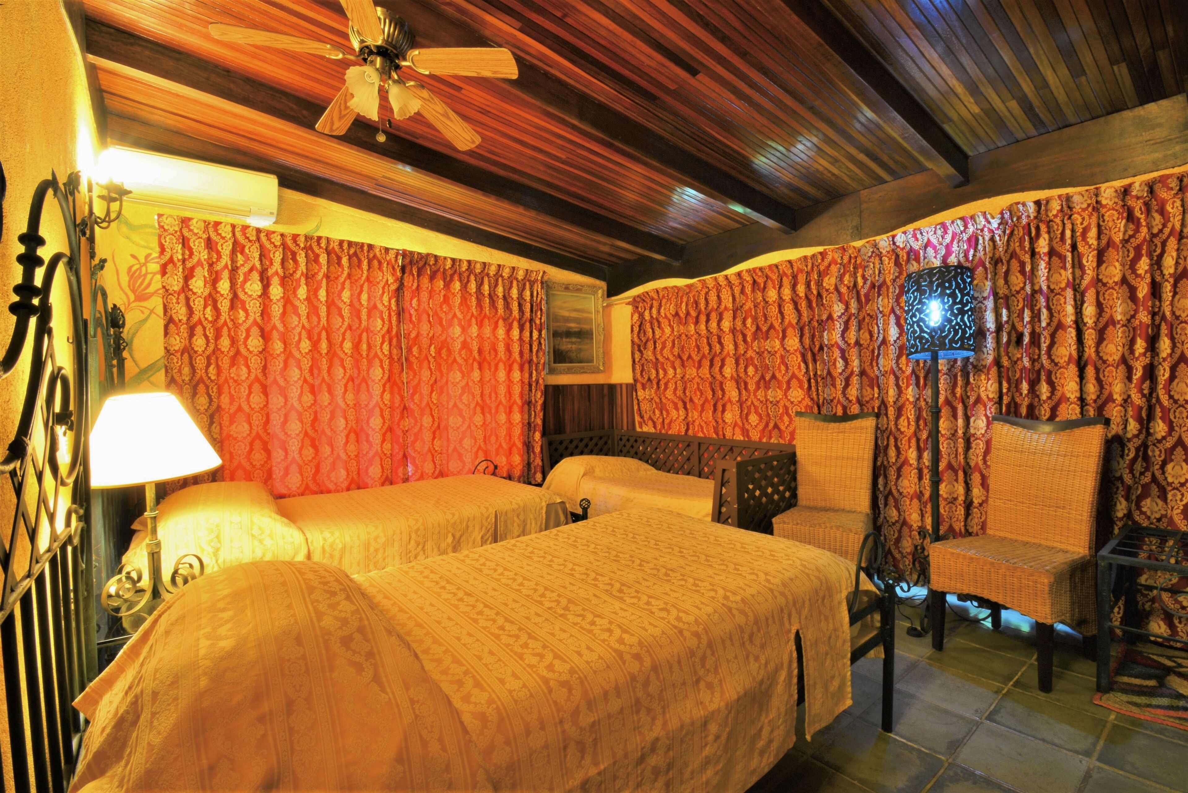 A room with two beds, a ceiling fan, patterned curtains, a lamp, and a chair.