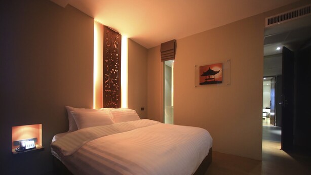 A modern hotel room with a large bed, a bedside table with a lamp, a decorative wall panel, and a painting of a pagoda. - Silom One (Bangkok)