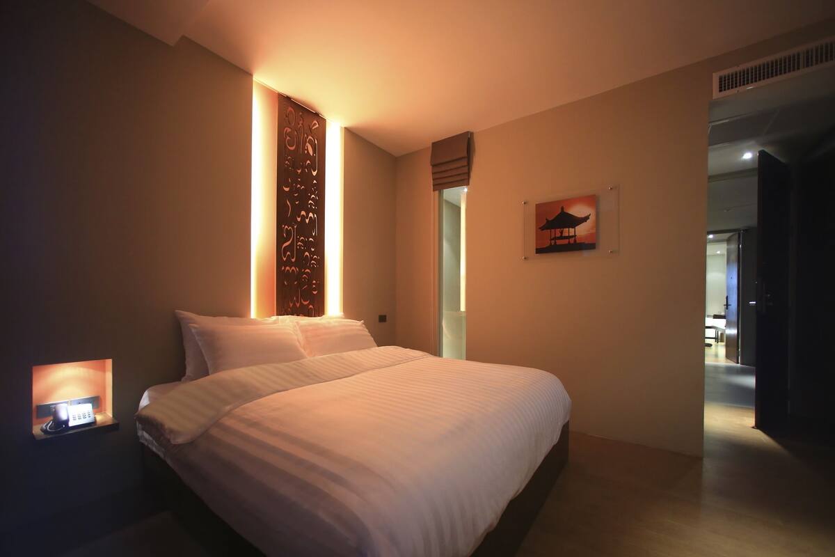 A modern hotel room with a large bed, a bedside table with a lamp, a decorative wall panel, and a painting of a pagoda.