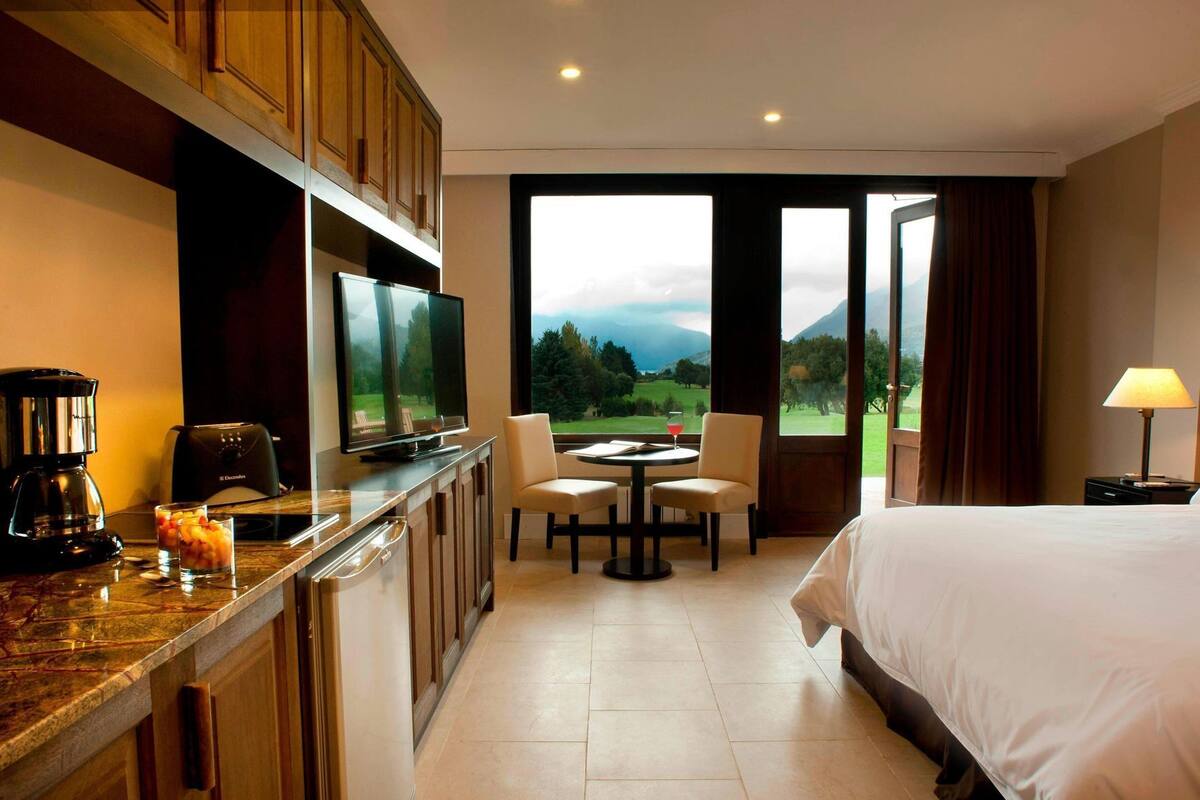 Habitación de hotel con cama, televisión, cafetera, mesa de comedor con sillas y vistas al exterior.