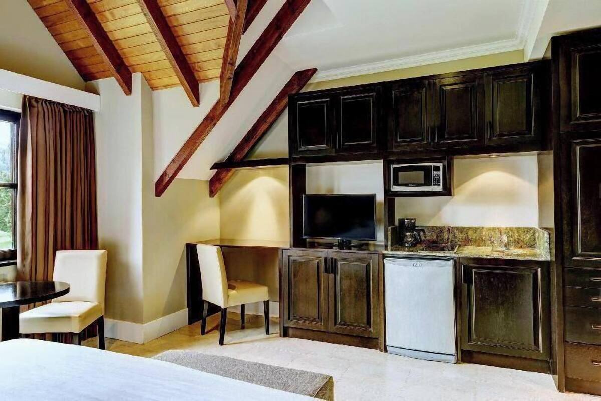 Cocina compacta con muebles oscuros de madera, microondas y un refrigerador pequeño. Hay una zona de comedor con mesa y sillas.
