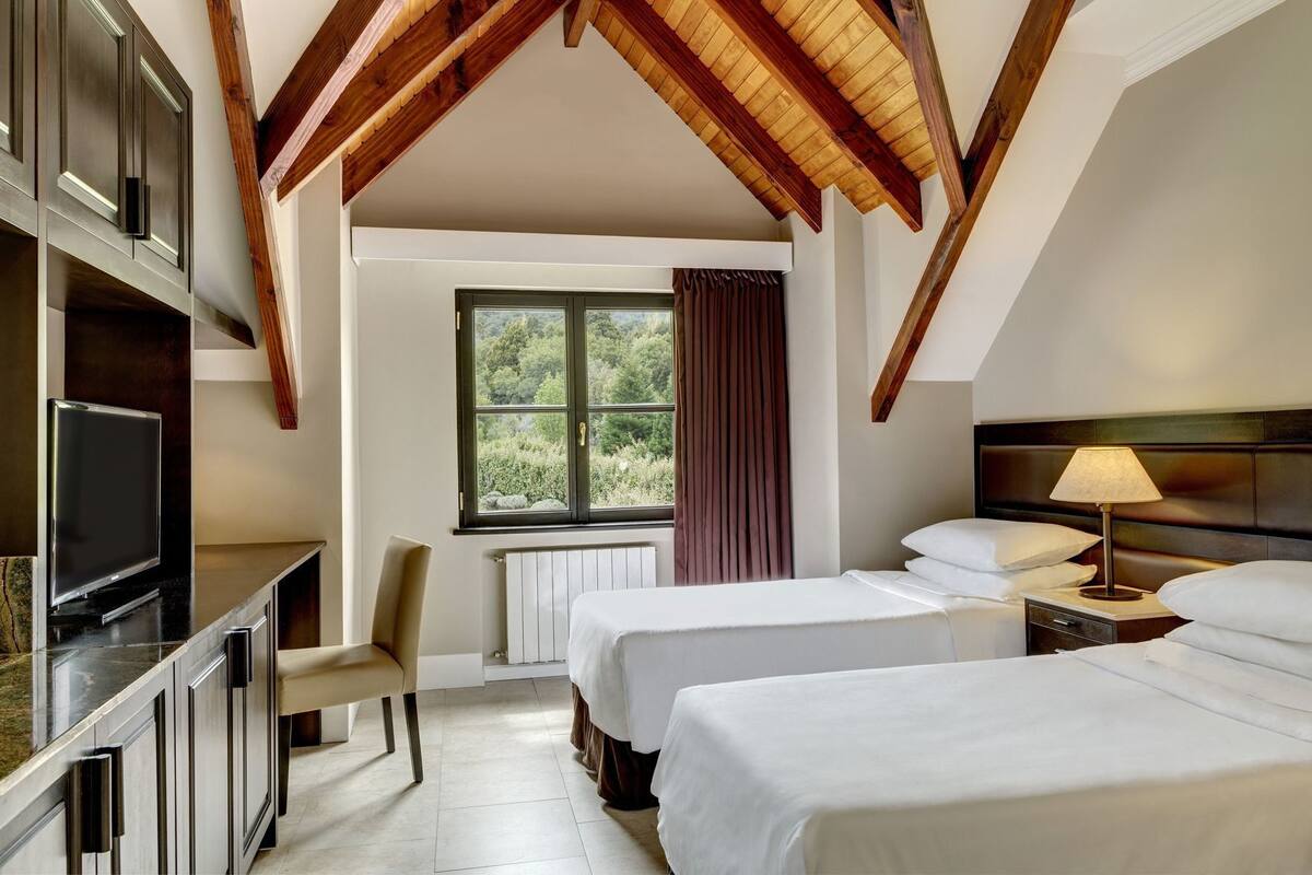 Habitación de hotel con televisión, escritorio, silla, dos camas, ventana con cortinas y lámpara.