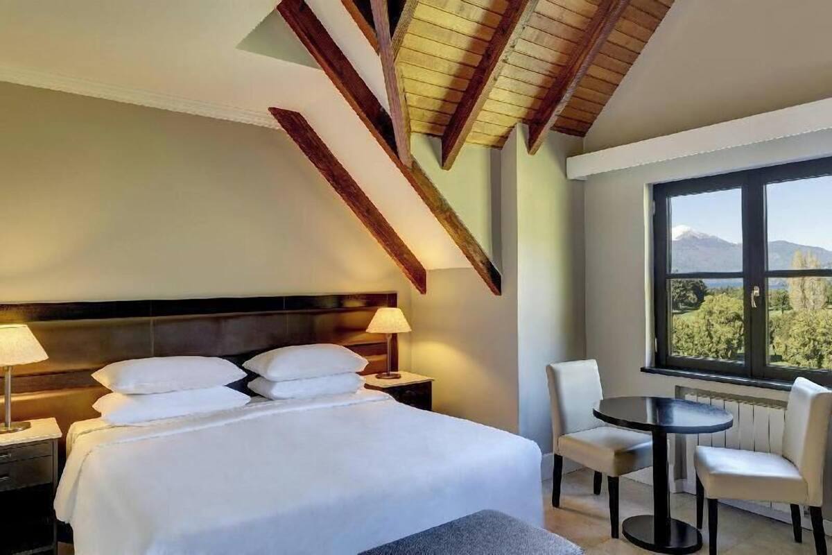 Habitación de hotel con una cama grande, mesitas de noche, una silla, una pequeña mesa y una ventana con vistas a árboles y montañas.