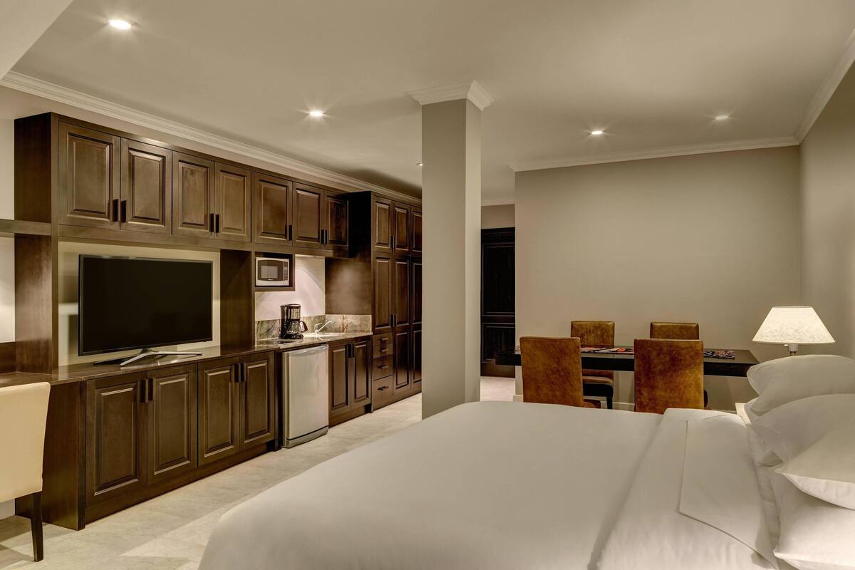 Una habitación de hotel moderna con una cama grande, televisor, kitchenette y escritorio con lámpara.