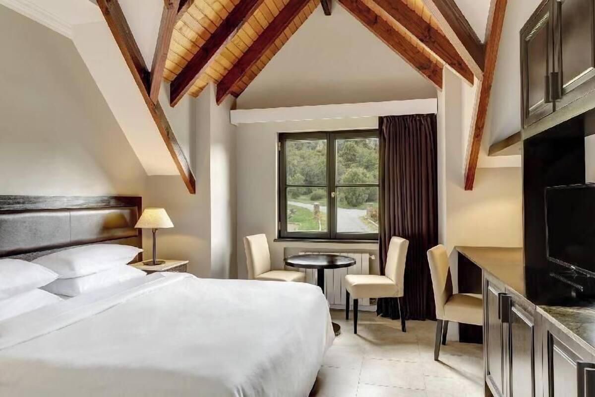 Habitación de hotel con cama, escritorio, silla, televisor y ventana con cortinas.