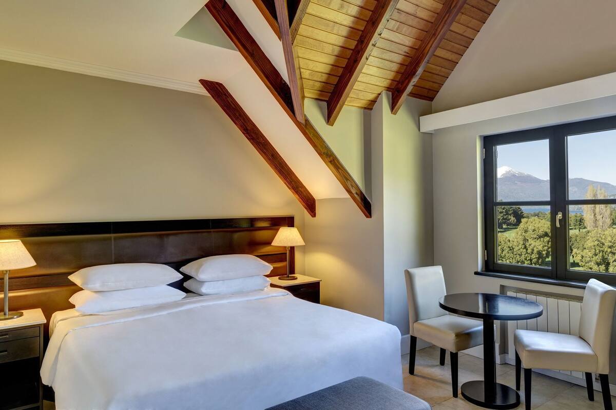 Una habitación de hotel con cama, lámparas de noche, una mesa redonda, dos sillas y un ventanal con vista a la montaña.